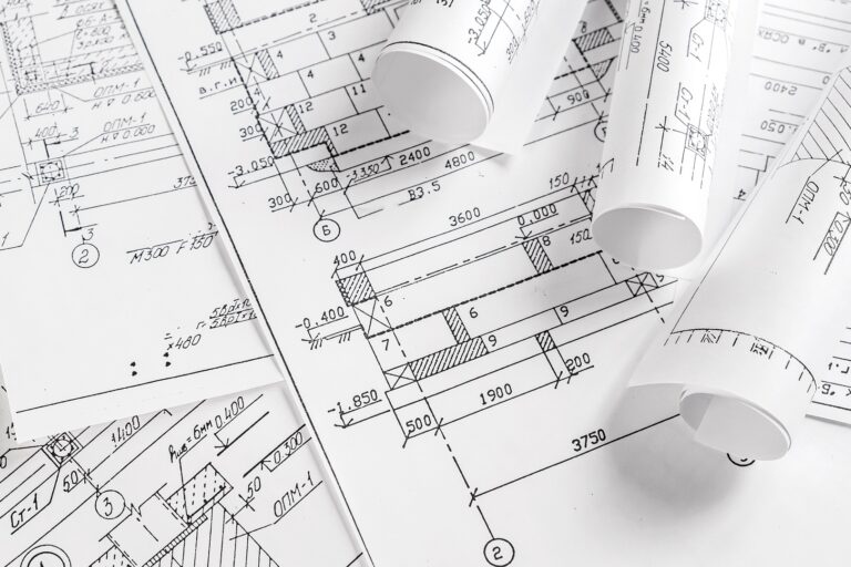 Drafting Services Several architectural blueprints with technical drawings and measurements, some flat and some rolled up, laid out on a white surface, showcasing professional Drafting Services.