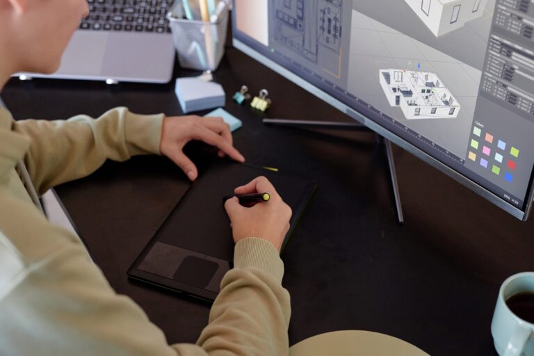 CAD Conversion Person using a digital drawing tablet while working on 3D architectural design software displayed on a computer monitor at a desk, showcasing advanced drafting services.