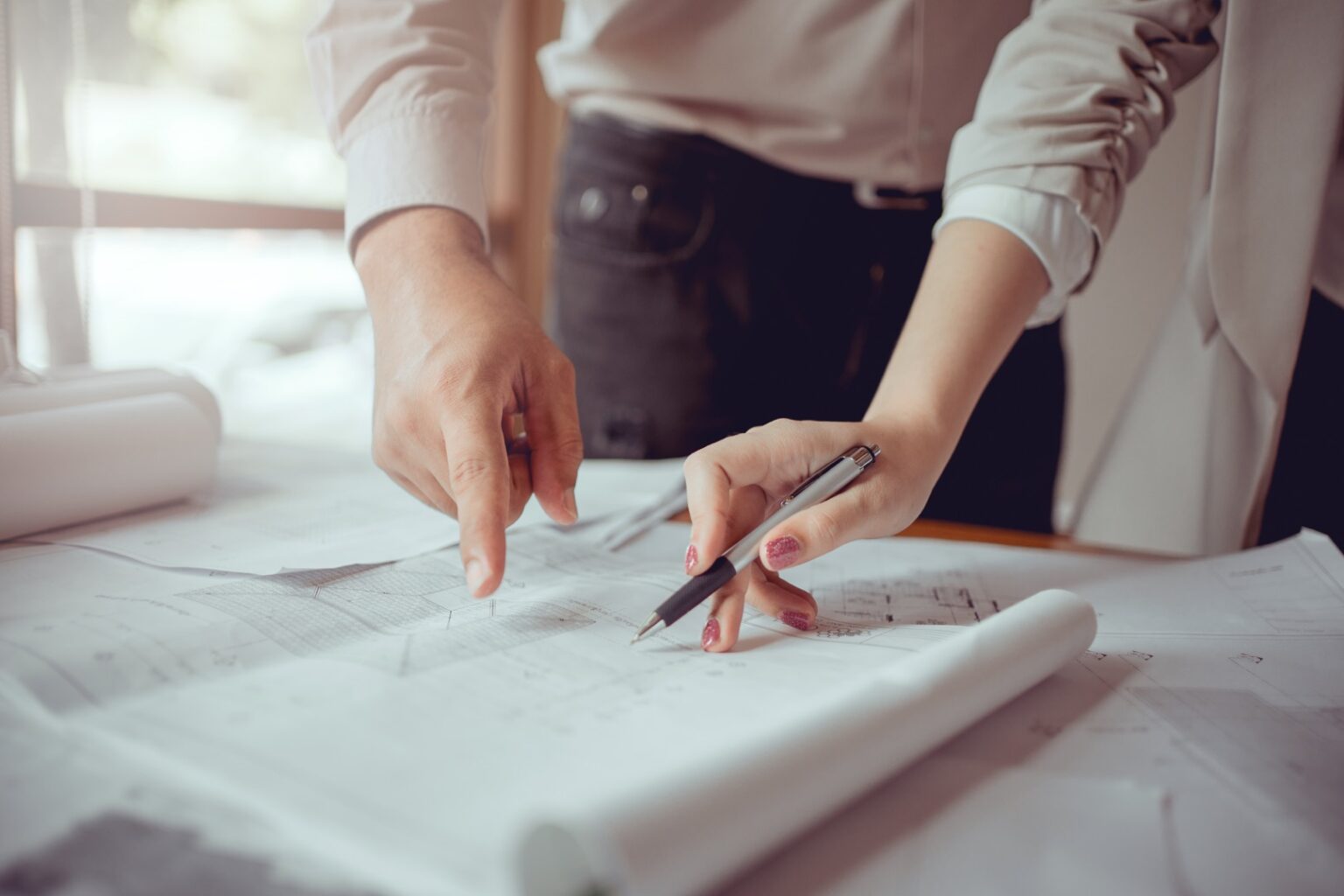 Two people standing and pointing at architectural blueprints on a table, one holding a pen and the other gesturing with a finger, showcasing detailed planning with drafting services and rolled paperwork nearby.