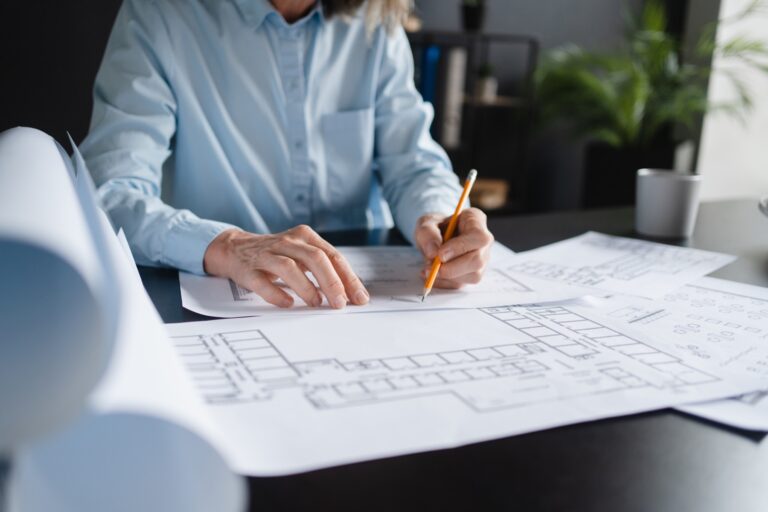 A person in a blue shirt reviews and marks architectural blueprints with a pencil at a desk, showcasing professional drafting services, with rolled-up plans and a coffee cup nearby.